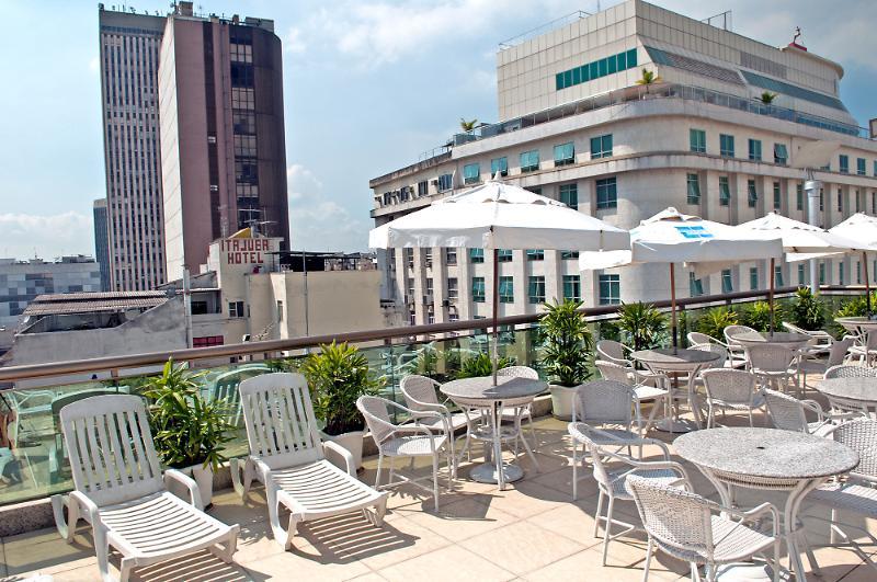 ריו דה ז'ניירו Hotel Atlantico Business Centro מראה חיצוני תמונה The photo shows a rooftop area with several white lounge chairs and circular tables. There are umbrellas providing shade over some of the seating. In the background, there are tall buildings, including a modern skyscraper with a distinct architectura